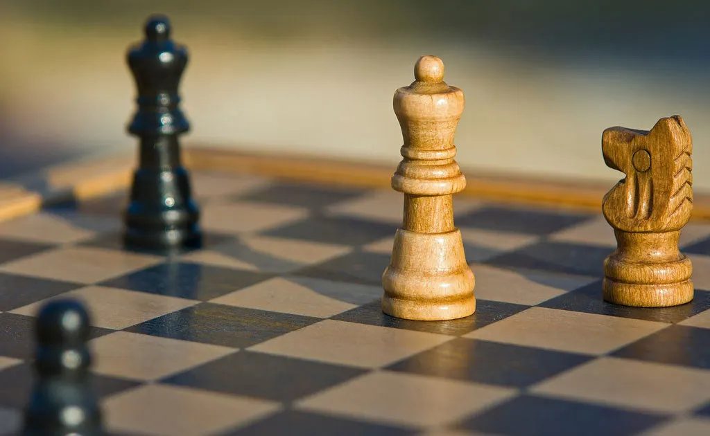 Chess pieces arranged on a board, with wooden pieces in focus and dark pieces blurred in the background.