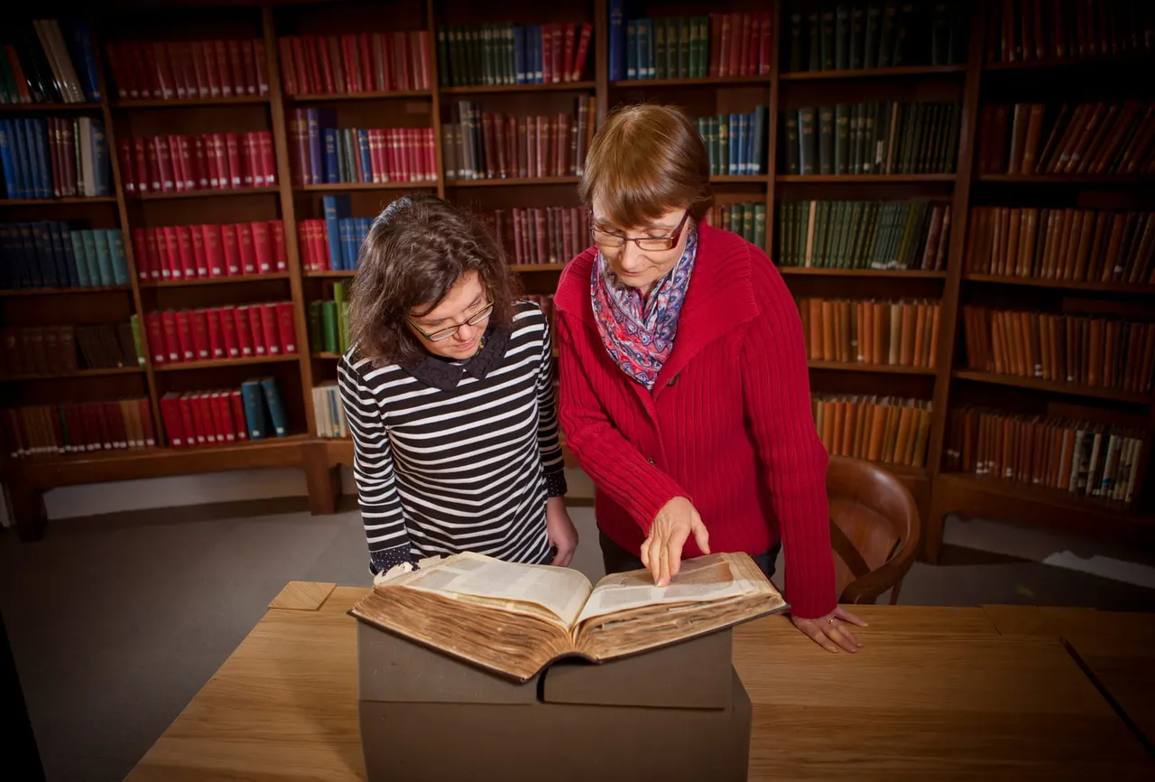 Two people examining an old book together at a table in a library with colourful bookshelves behind them.