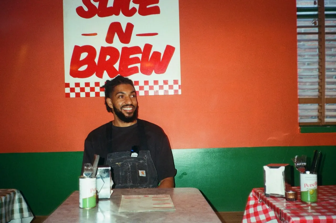 Man seated at a table in a casual dining venue with red and green walls and a "Slice 'n' Brew" sign behind him.