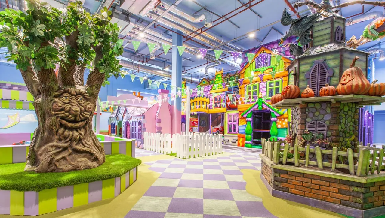 Colourful indoor soft play area with themed buildings, a large tree sculpture, and checkered floor in bright greens and purples.