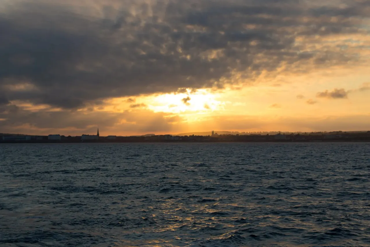 Sunset over water with distant coastal town silhouetted against golden sky and dramatic clouds.