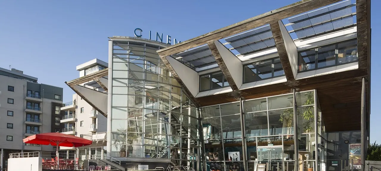 Modern cinema building with glass frontage, red umbrellas on outdoor seating area, and "CINEMA" sign visible on roofline.