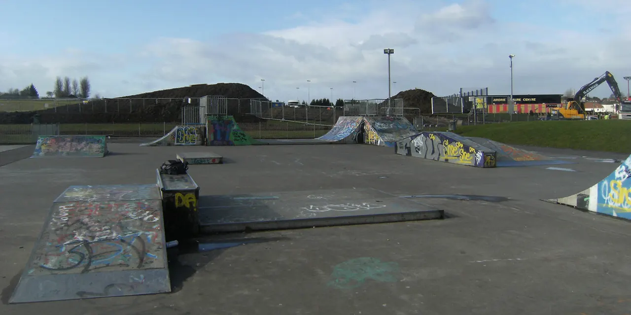 Outdoor skate park with graffitied ramps and rails on asphalt, surrounded by fencing and floodlights.