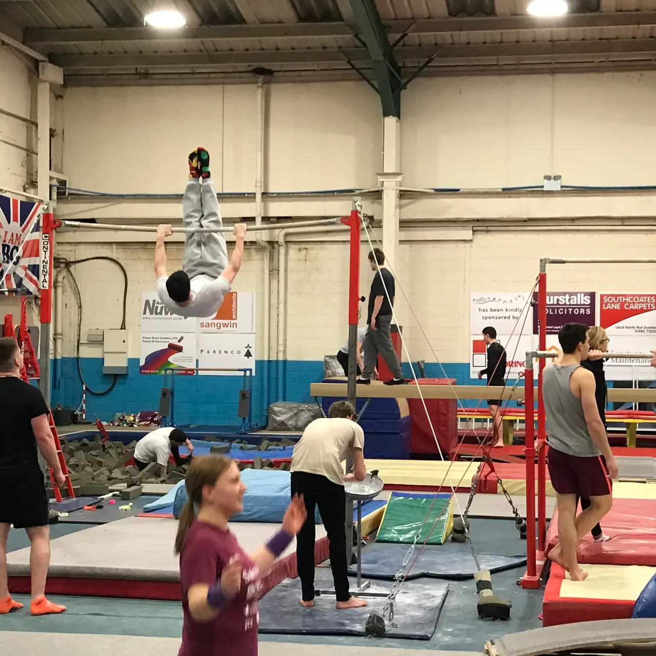 Multiple young gymnasts training on apparatus in an indoor gymnastics hall, with one performing a backflip on a bar.