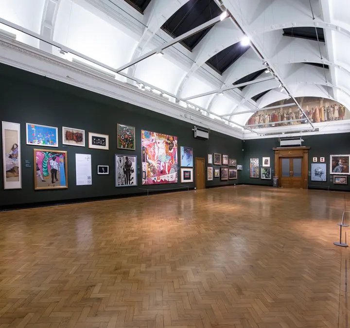 Spacious art gallery interior with dark green walls displaying various artworks, wooden herringbone floor, and skylit ceiling with white metal framework.