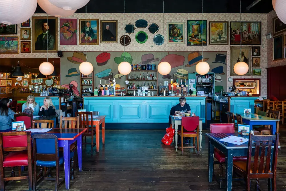 Colourful vintage-style bar interior with bright blue counter, mismatched furniture, hanging globe lights, and eclectic wall art including paintings and decorative hats.