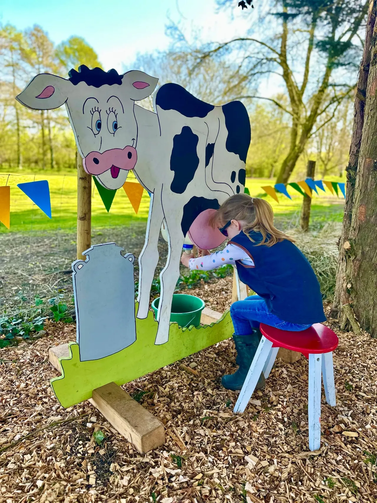 Child in pink cap playing with a milk bottle prop at a cartoon cow cutout in a woodland play area.