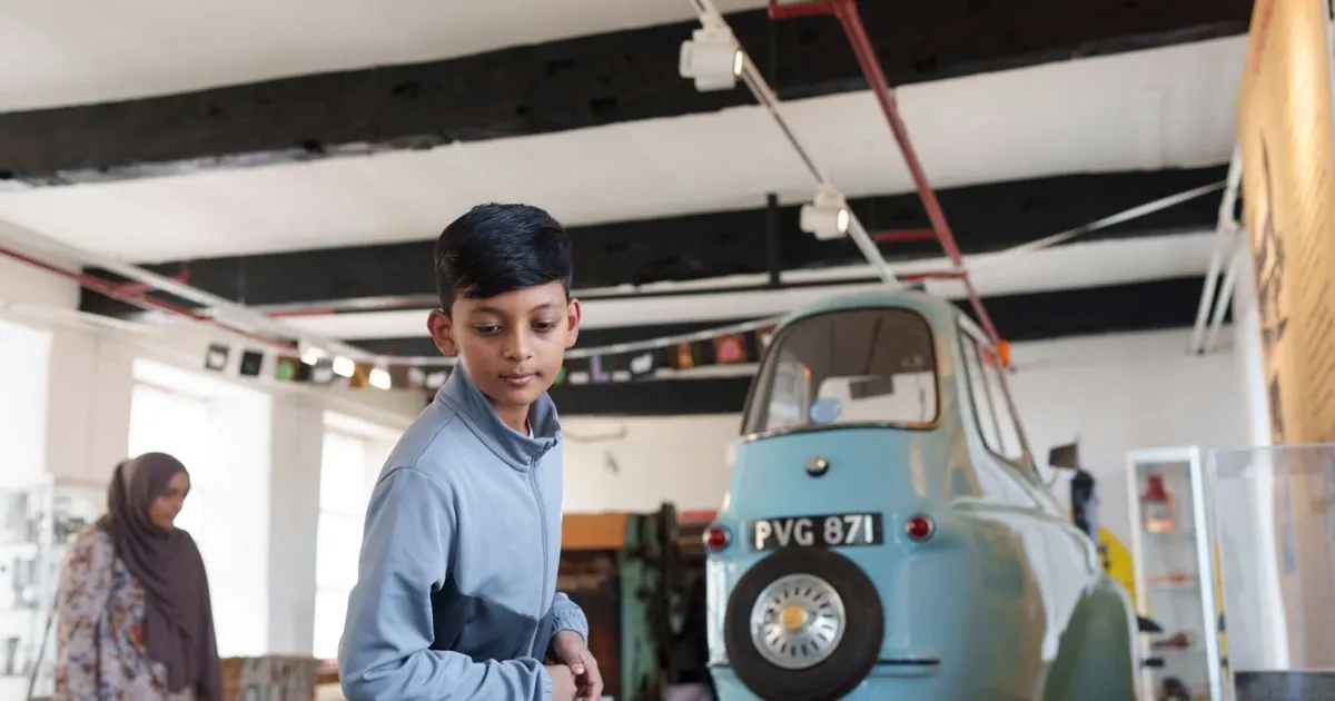 Boy in blue shirt stands beside a turquoise vintage car in an industrial museum exhibition space.