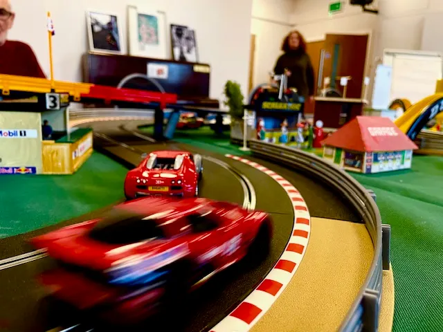 Toy racing cars race on a slot car track in an indoor play area with families nearby.