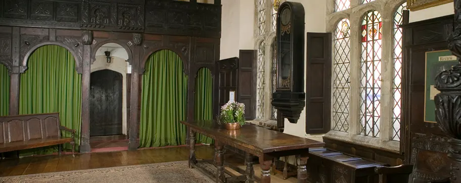 Historic interior hall with wooden panelling, green curtained alcoves, period furniture, and leaded windows.