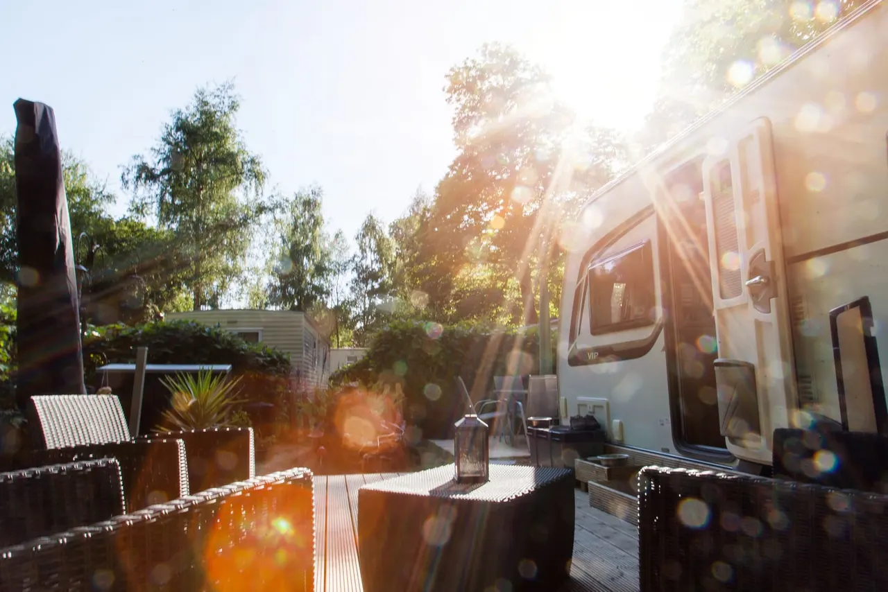 Glamping site with a silver caravan and decked outdoor seating area surrounded by trees at sunset.