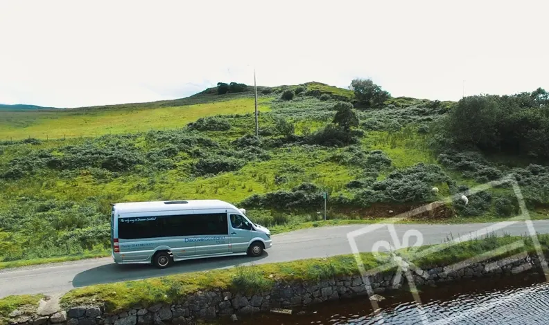 A minibus parked on a rural road with green hillside and water in the foreground.