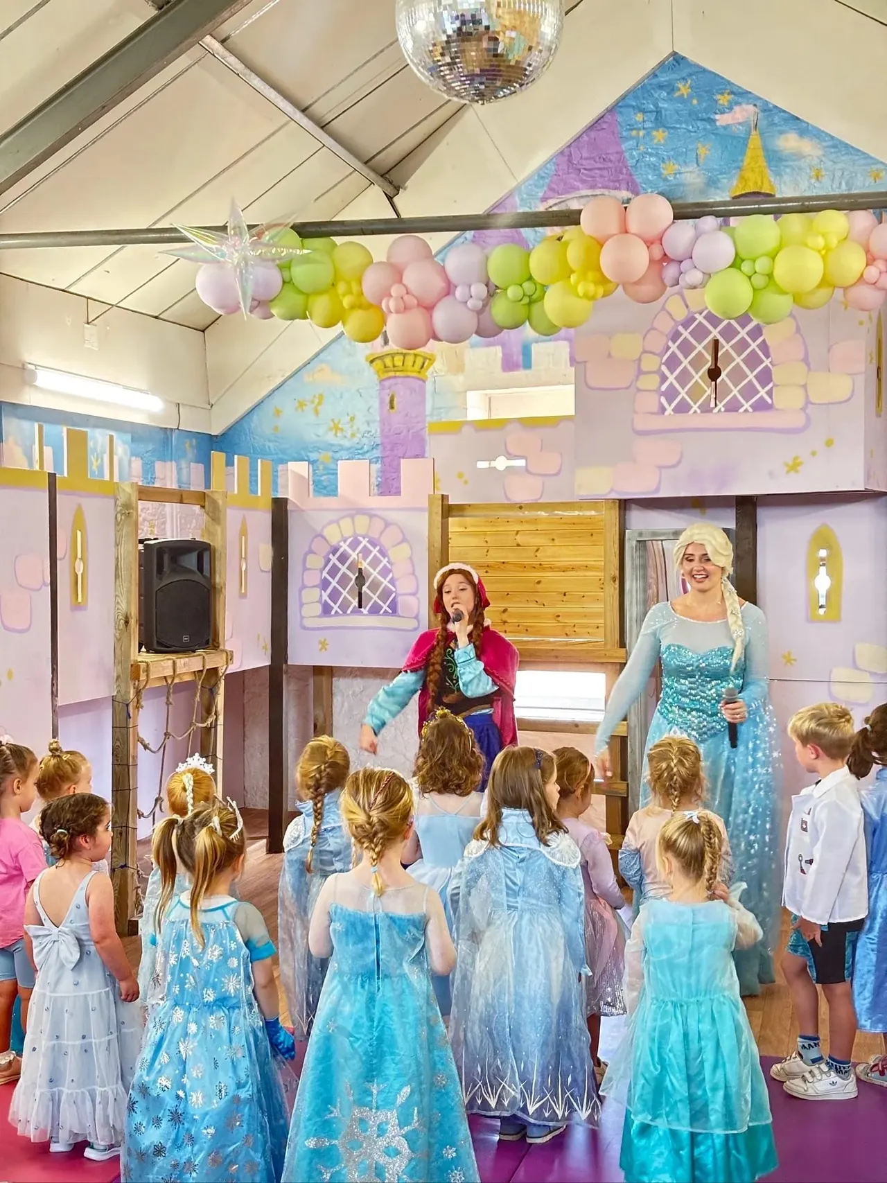 Children in Disney-themed costumes gathered around two costumed characters in a decorated castle-themed indoor play space.
