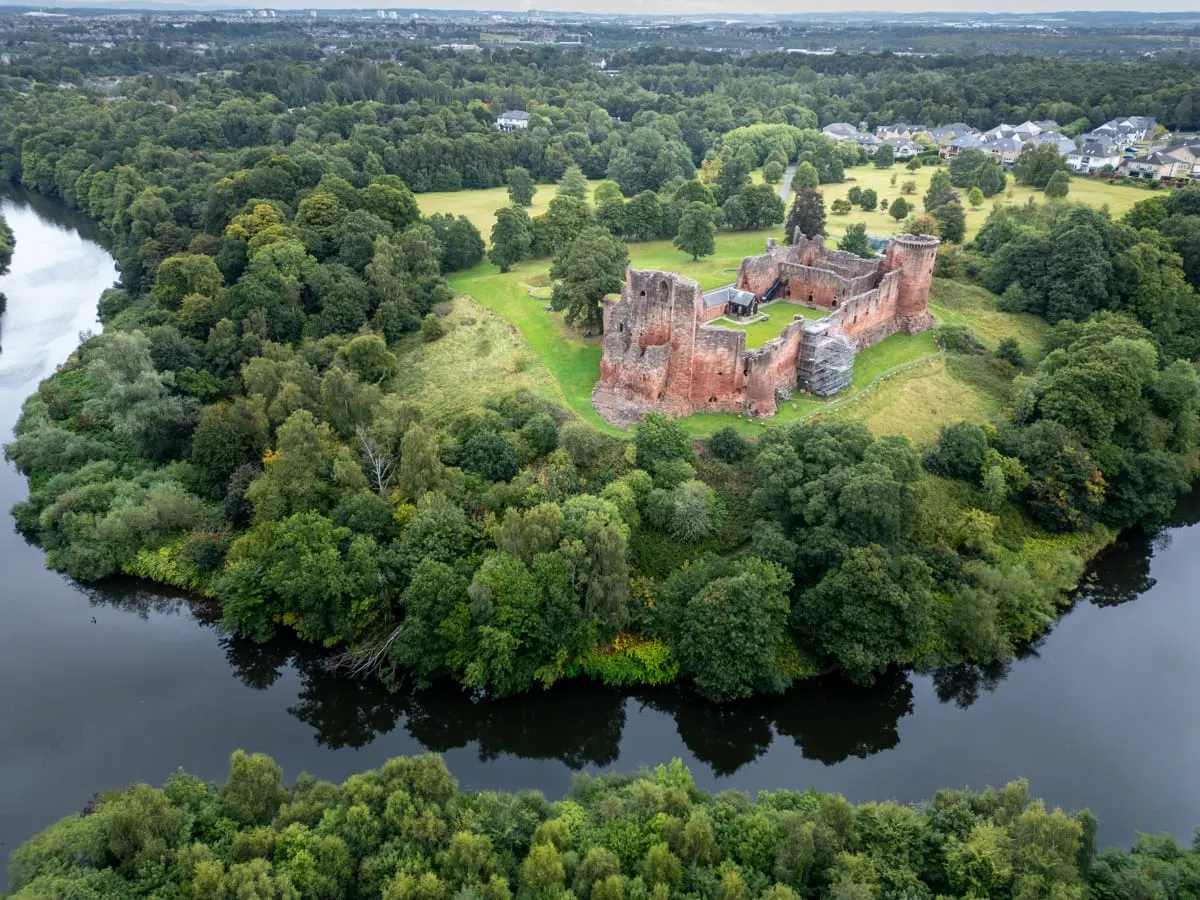 Aerial view of a red sandstone castle ruins on a peninsular surrounded by river, woodland and green fields, with distant houses and town beyond.