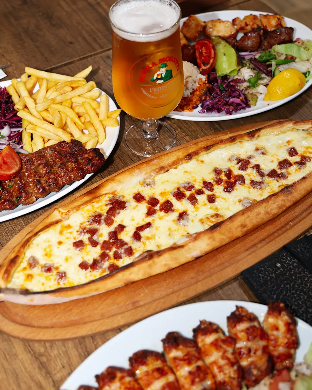 Table spread with cheese and bacon flatbread, grilled meat kebabs, chips, salad, and beer on wooden surface.