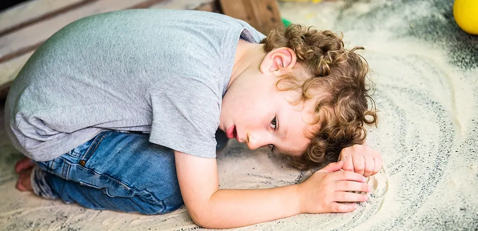 Young child with curly brown hair crouched down examining something on the ground outdoors.