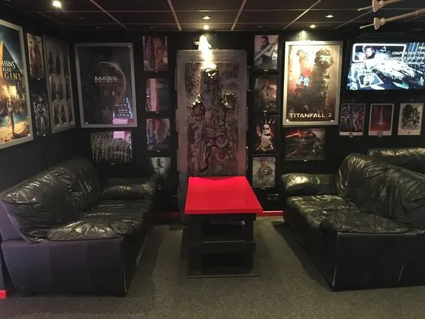 Gaming lounge interior with black leather seating, red table, and video game posters on dark walls.