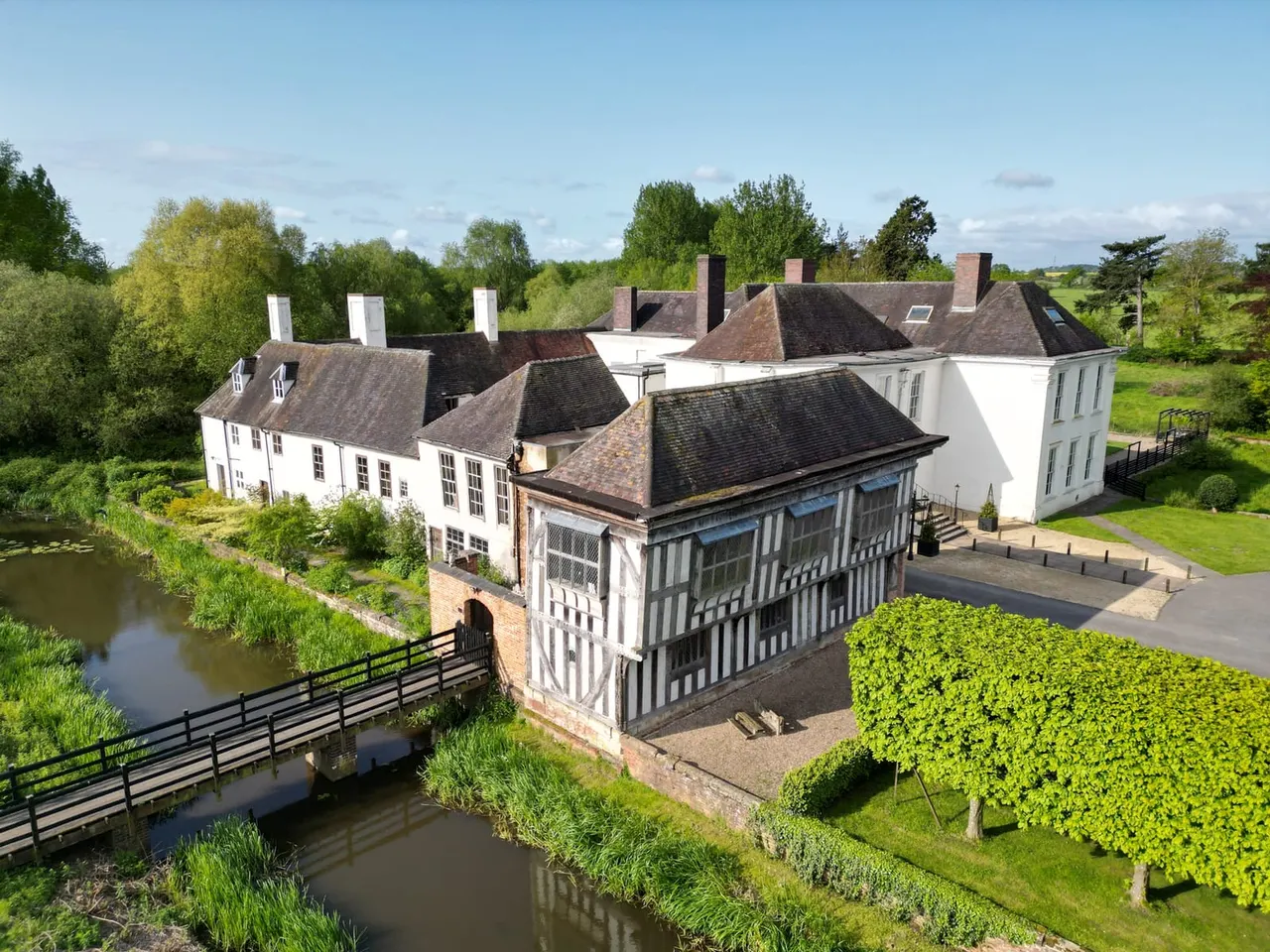 Historic half-timbered manor house with multiple chimneys beside a moat and bridge, set in parkland with manicured gardens and surrounding trees.