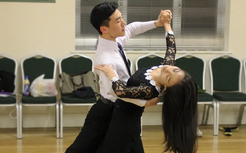 Male and female dancers performing a ballroom dance move in a studio with empty chairs in the background.