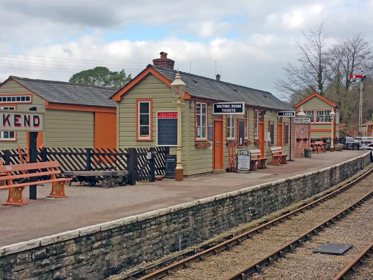 Historic railway station buildings with orange and cream painted wooden structures, railway tracks in foreground, and benches on platform.