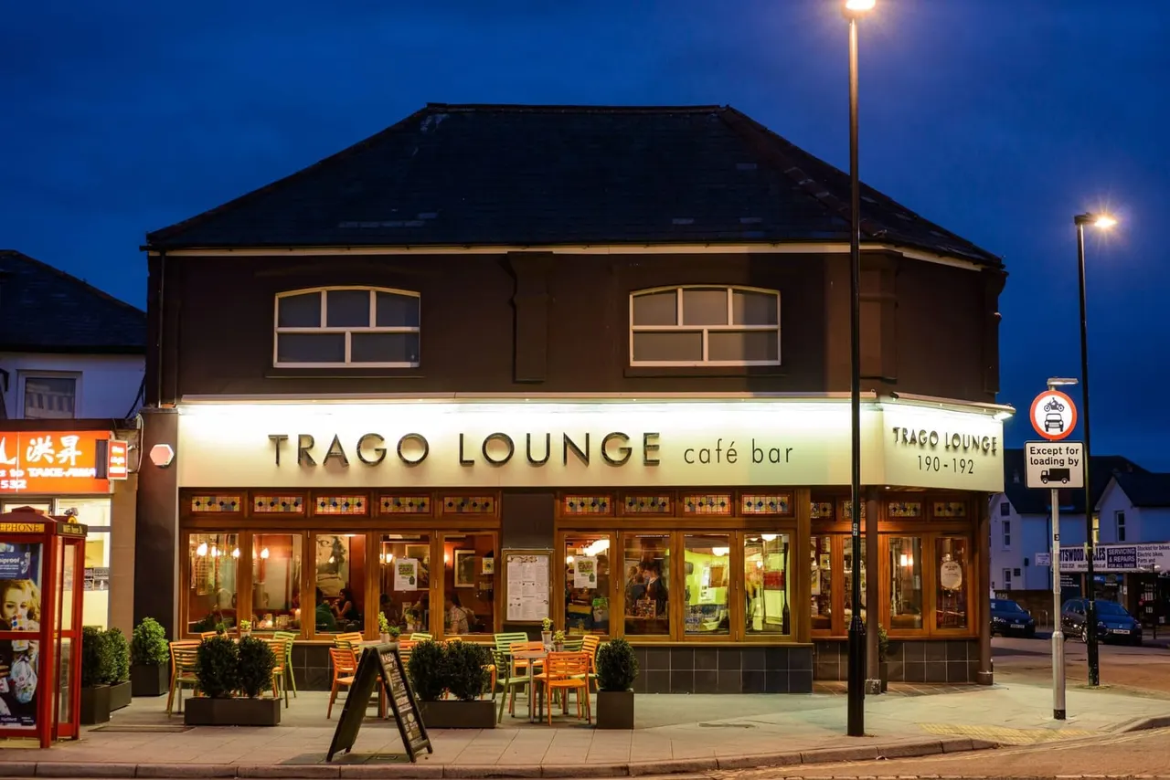 Trago Lounge café bar storefront at dusk with lit windows, outdoor seating, and people visible inside.