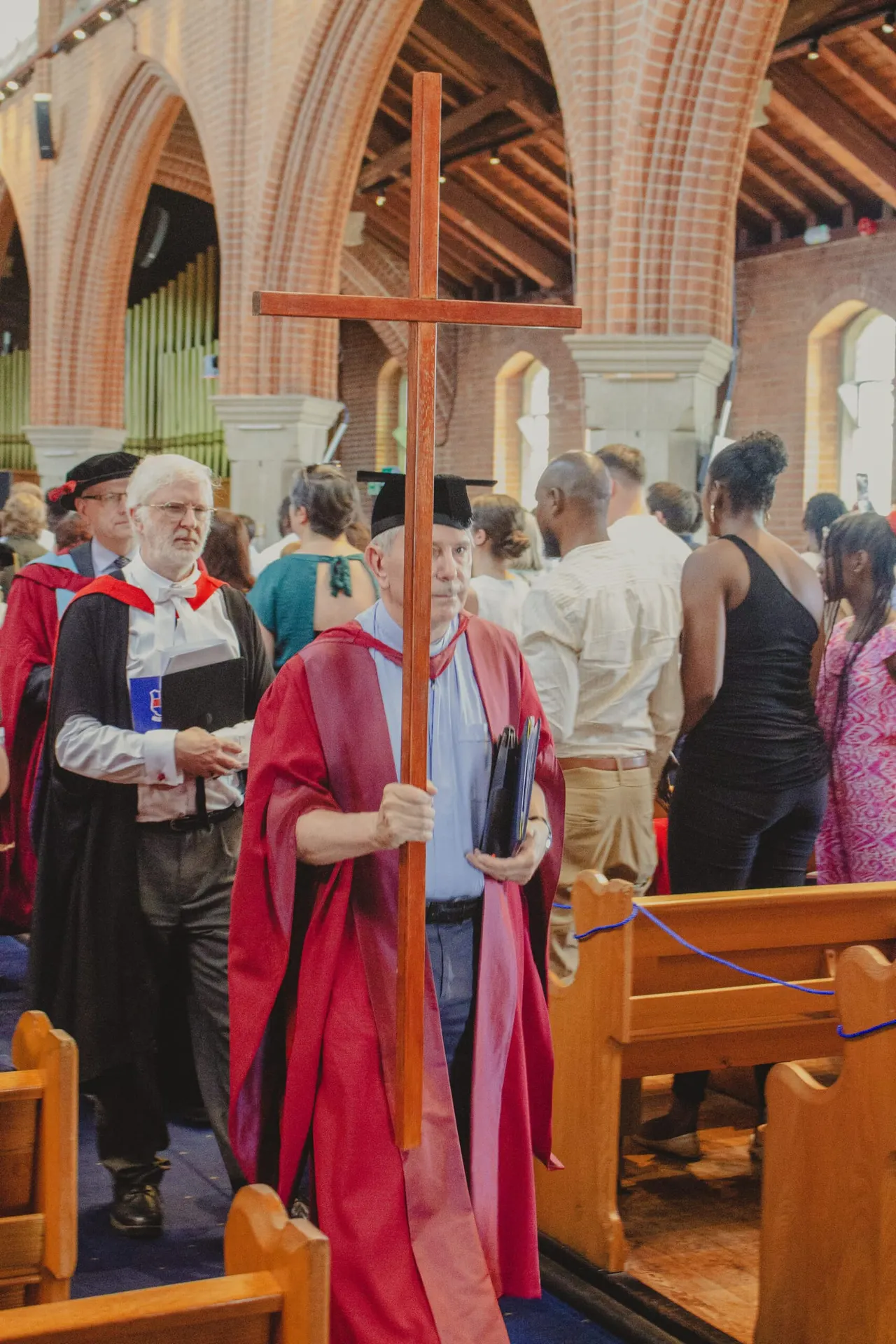 Clergy member in red vestments carrying a wooden cross during a church service with congregation gathered inside a brick chapel.
