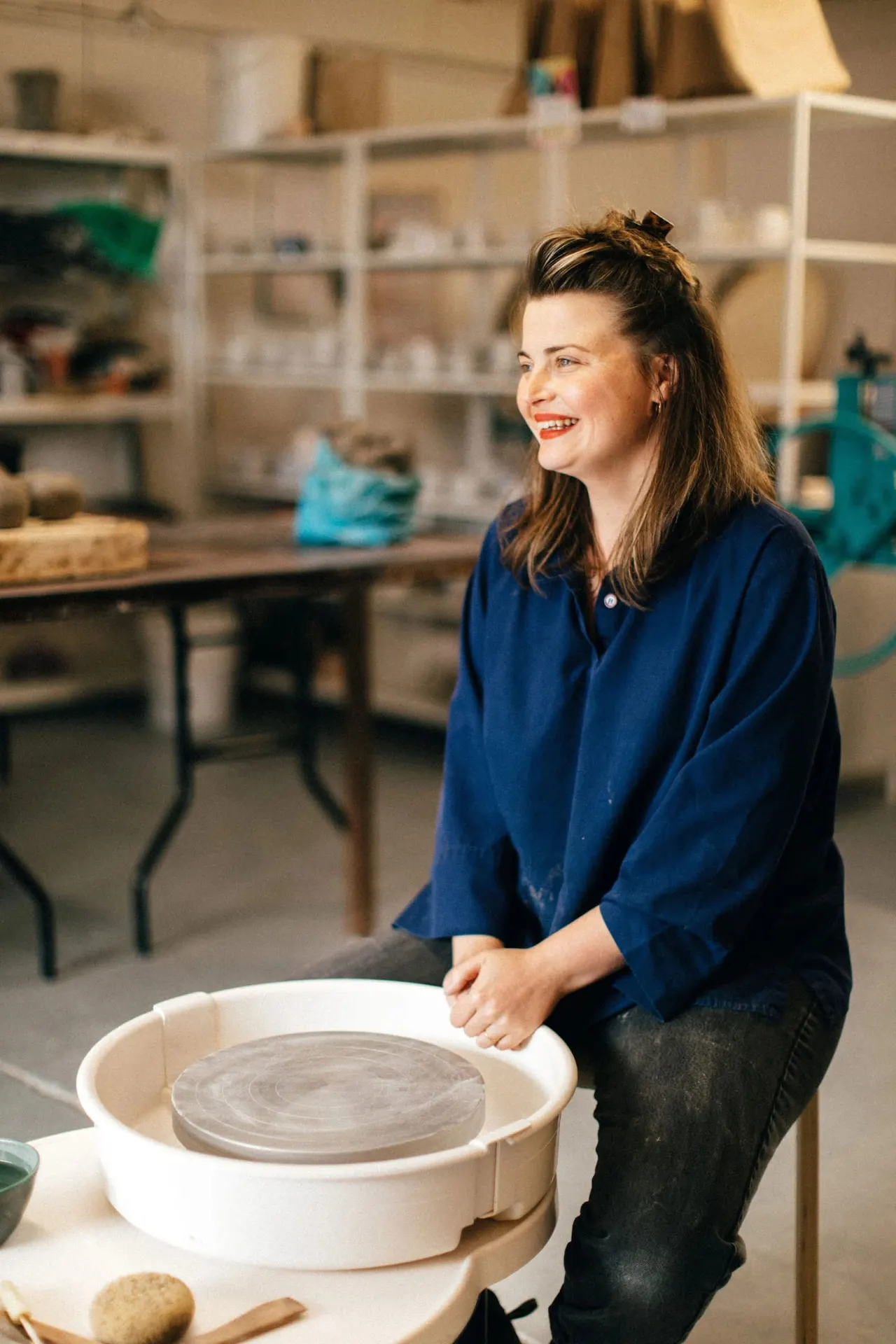 Woman in blue shirt working at a pottery wheel in a studio with shelving and materials in the background.