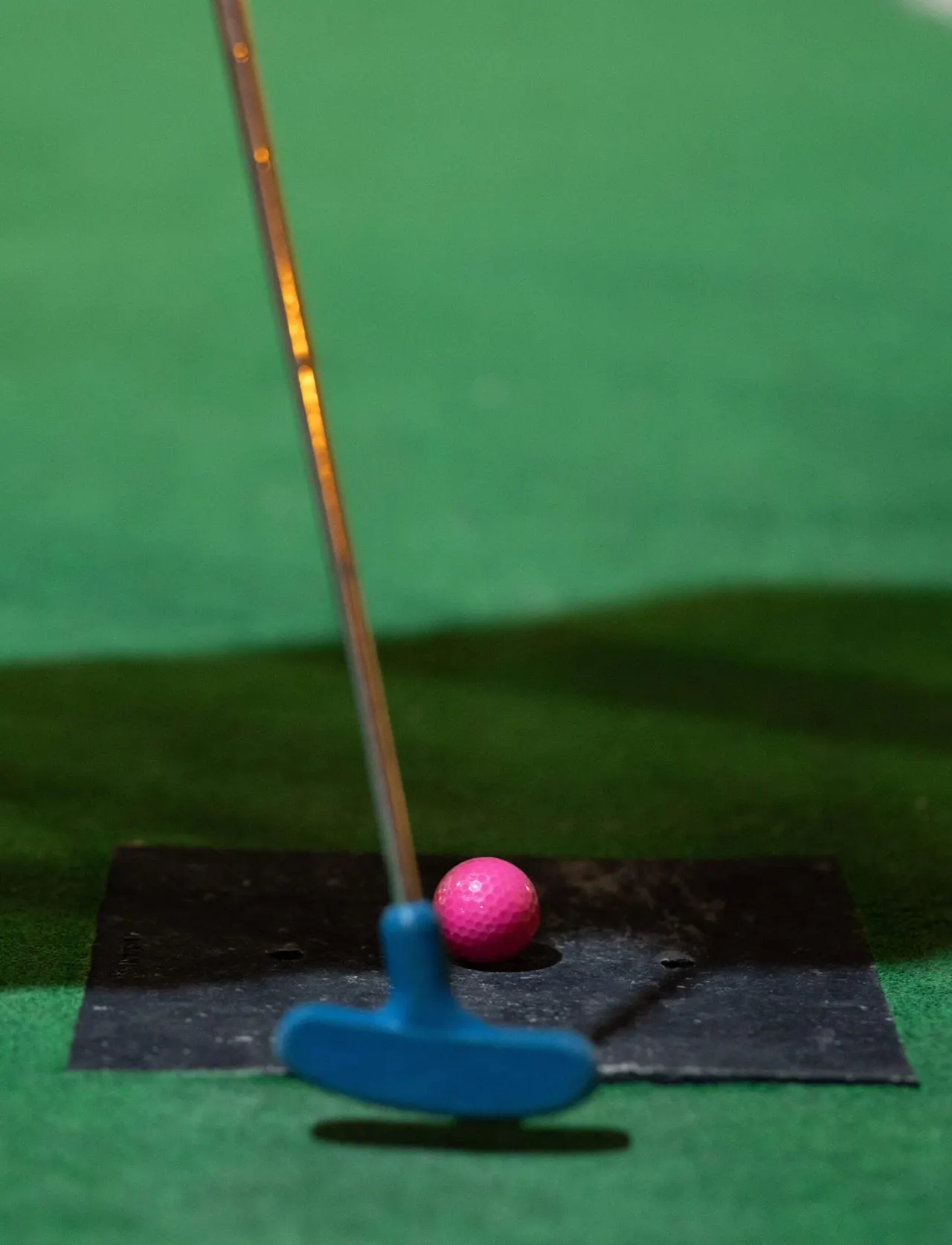 A blue putter with a pink golf ball on the putting mat at an indoor mini golf course.