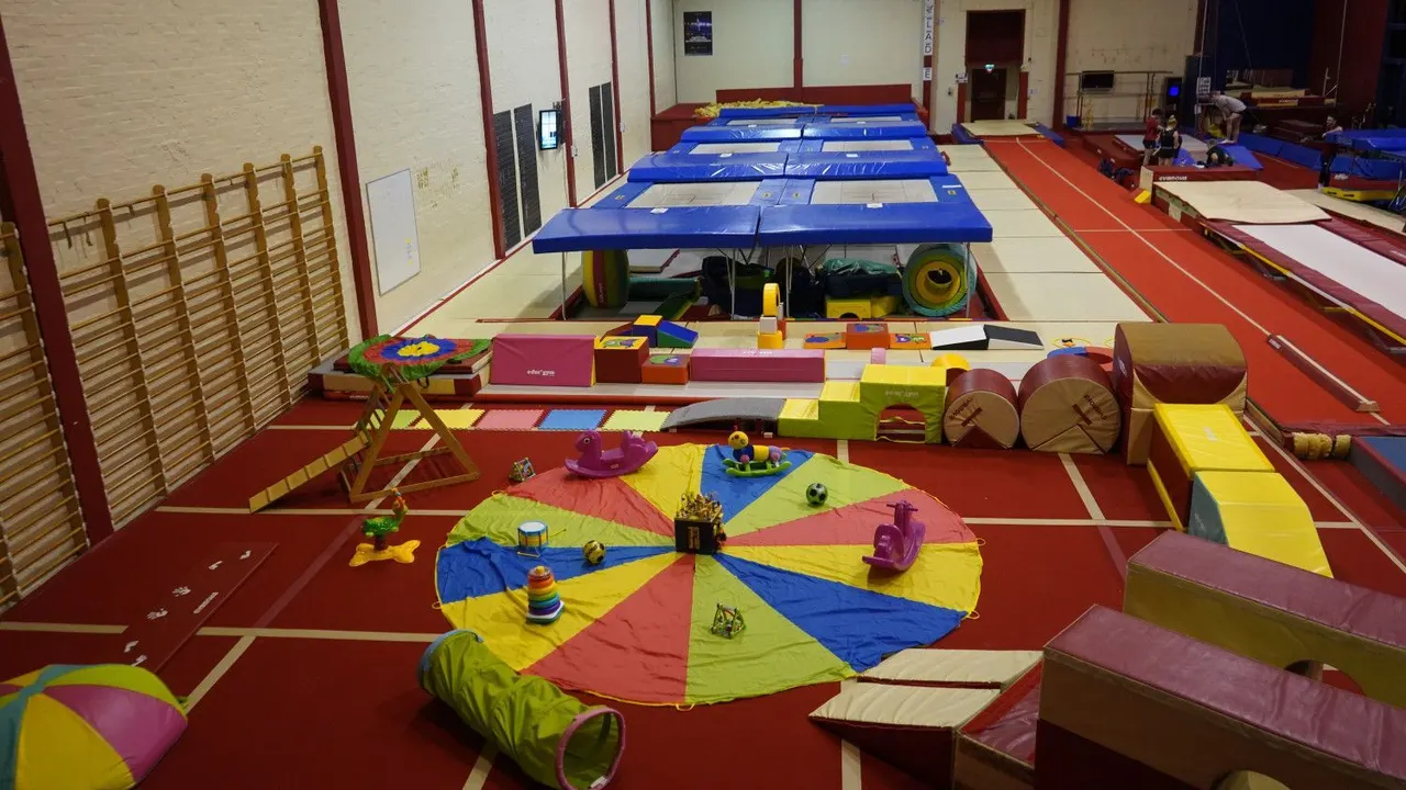 Indoor gymnastics hall with colourful play equipment, trampolines, soft mats and climbing apparatus arranged across a sprung floor.