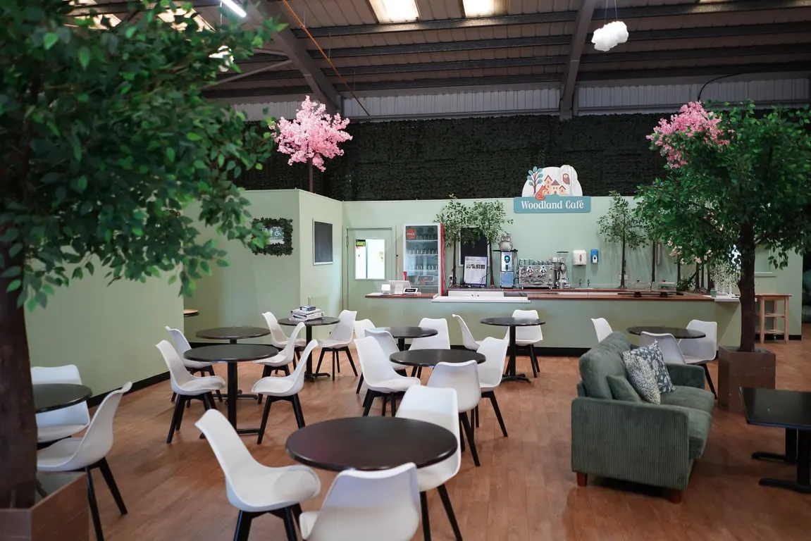Interior of a spacious indoor café with tables, white chairs, a service counter, potted plants, and decorative pink flowers hanging from the ceiling.