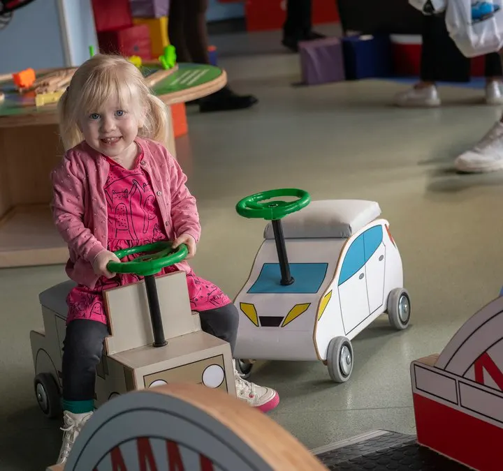 A young child in a pink jacket rides a toy vehicle indoors at a play facility, smiling at the camera.