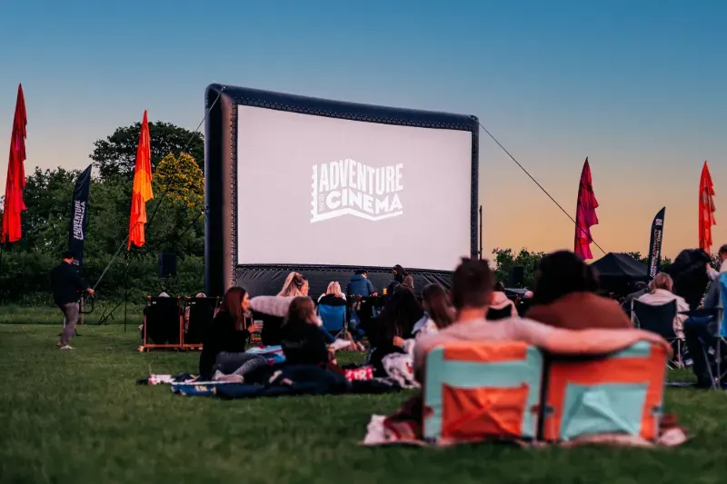 Outdoor cinema event at dusk with crowds seated on grass watching a large inflatable screen surrounded by colourful flags.
