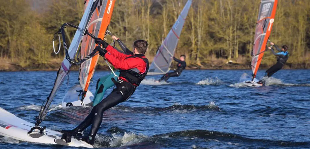 Windsurfers in wetsuits riding waves on a lake with coniferous forest in the background.