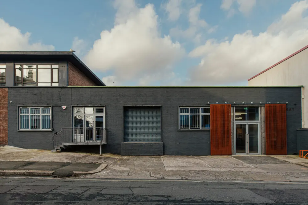 Modern single-storey building with dark brick facade, red metal doors, and windows along a quiet street.