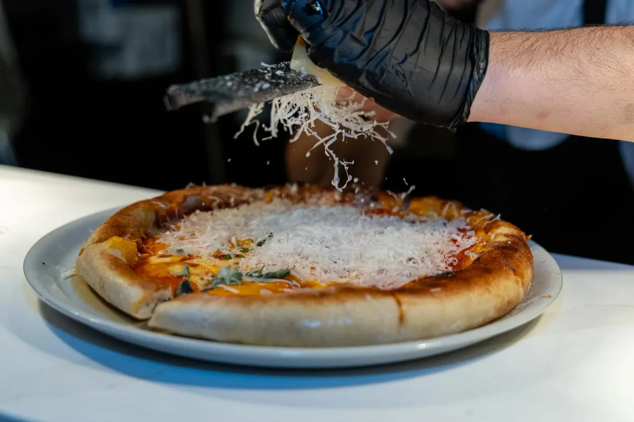 Person in black glove holding a small fish above a freshly made pizza on a plate.