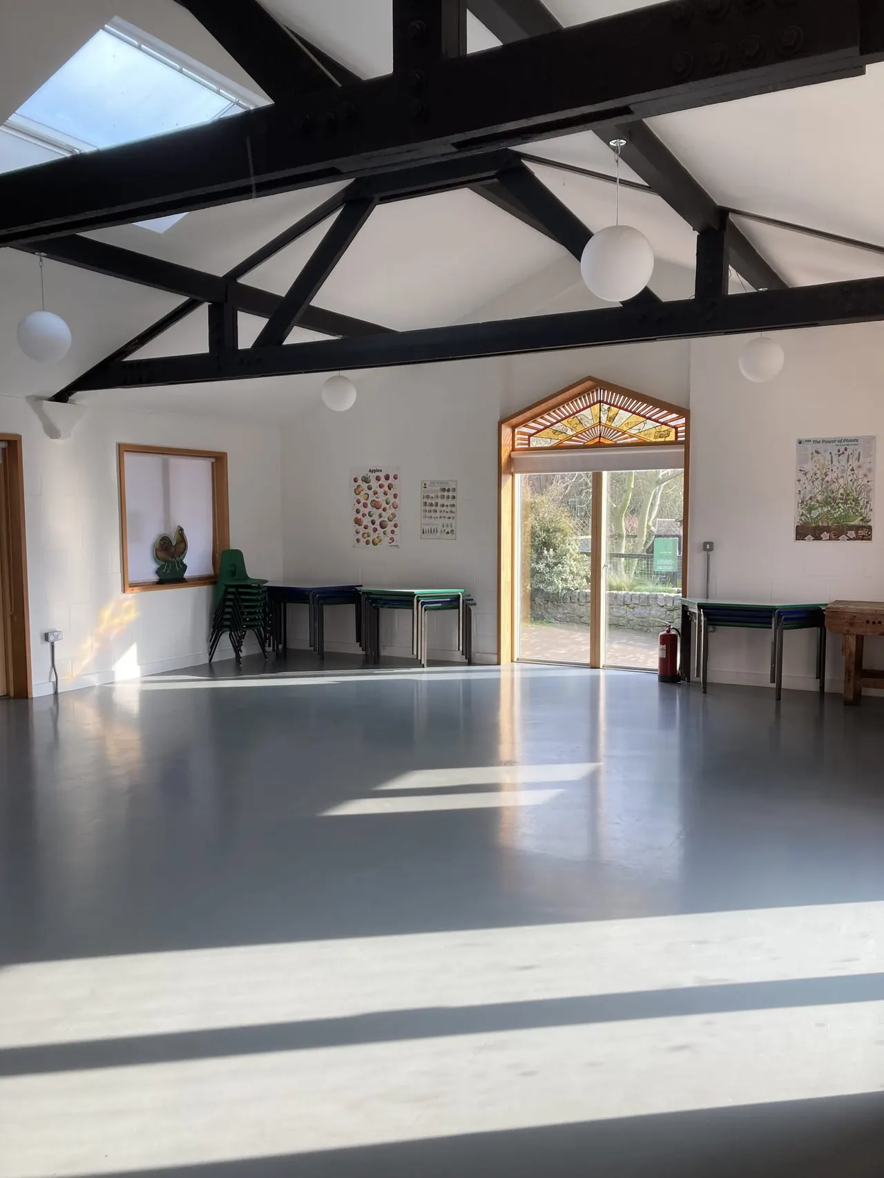 Spacious bright hall with polished grey floor, black exposed beams, skylights, stacked chairs and glass doors opening onto gardens.