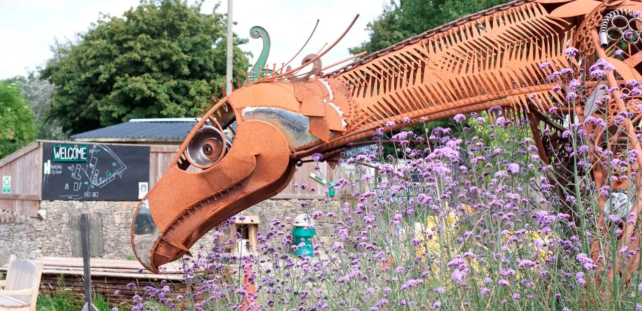 Large copper insect sculpture with purple flowers blooming in front, beside a welcome building and green trees.