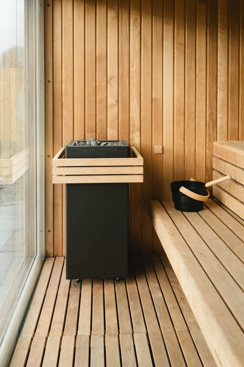 Interior of a wooden sauna with benches, a black stove with wooden surround, and a black ladle with wooden handle.