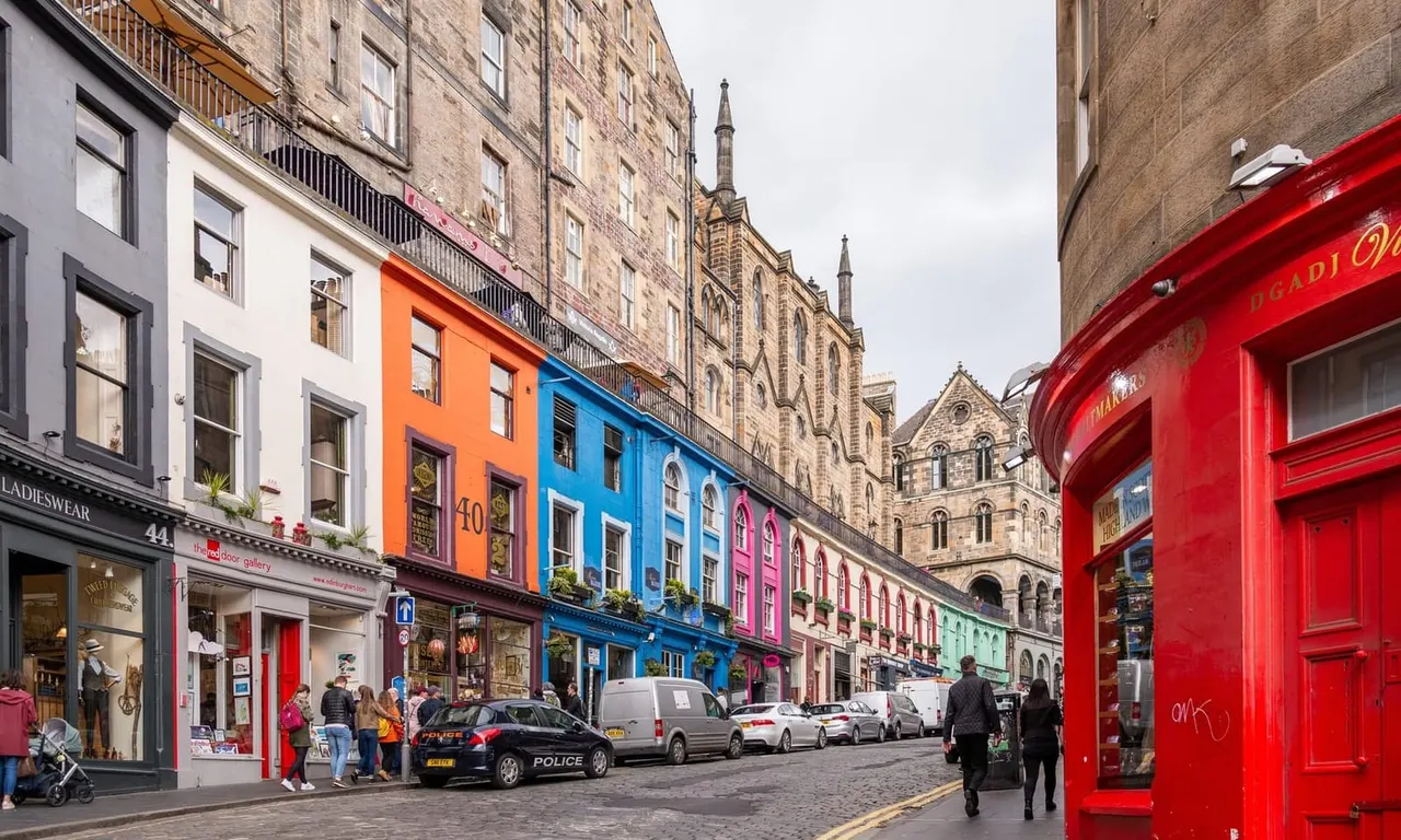 A cobbled street lined with colourful shop fronts and historic stone buildings, with pedestrians and parked vehicles.