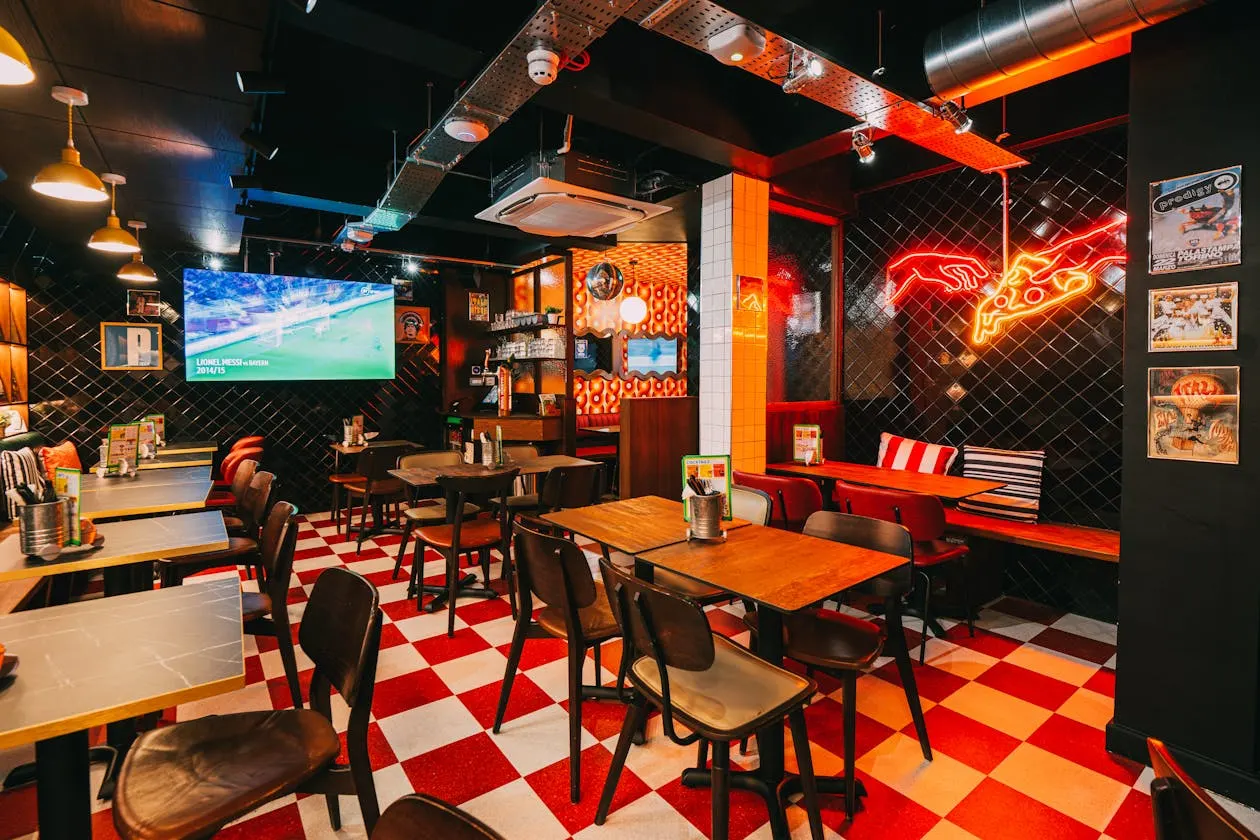 Interior of a pizza restaurant with red and white checkered floor, wooden tables, neon signage, and a TV screen showing sports.