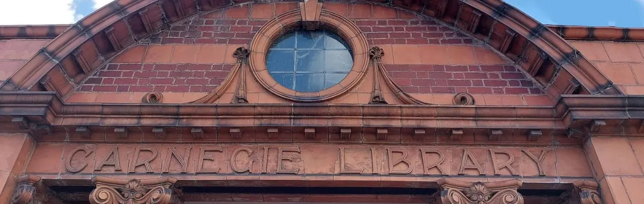 Red brick Victorian building façade with ornate circular window and "Carnegie Library" carved lettering.