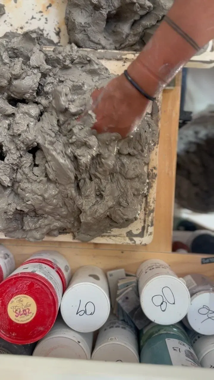 Person's hand working with wet clay on a pottery wheel, with paint containers visible below.