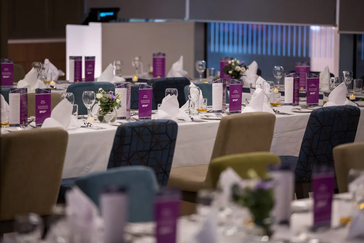 Formal banquet hall with white-dressed long tables, purple menu cards, white napkins, floral centrepieces, and place settings ready for a corporate event or gala dinner.