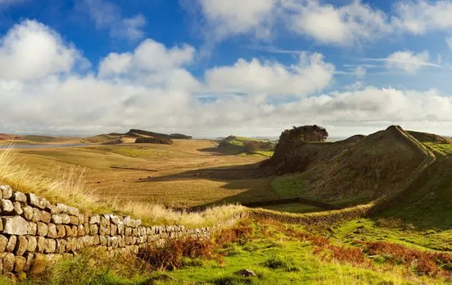 Rolling moorland with stone wall, grassy ridge and distant hills under blue sky with white clouds.