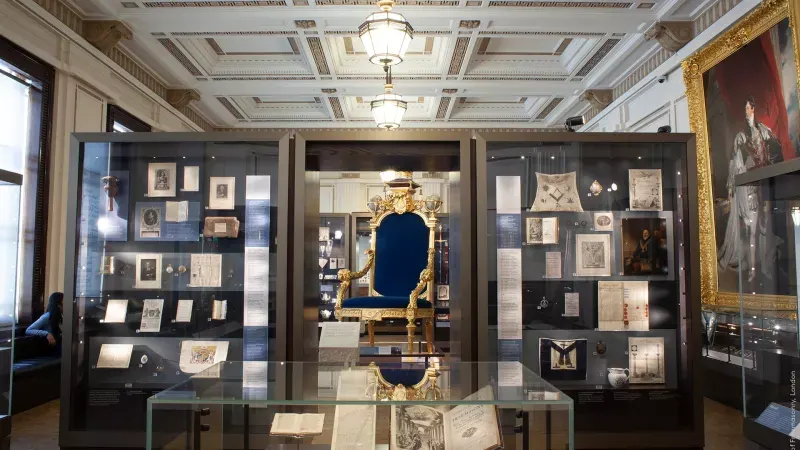 Museum display room with glass cases containing historical documents, artefacts and a gilded ornamental chair in the centre.