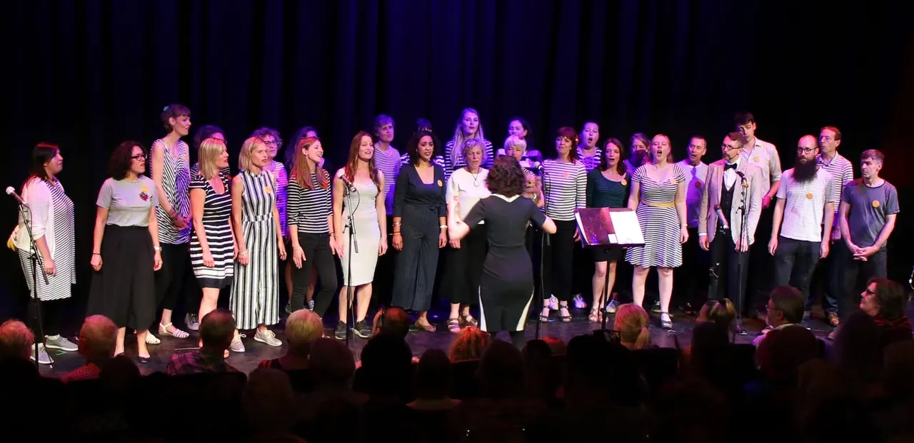 A large choir performs on stage under purple lighting, with many members wearing striped shirts, whilst an audience watches from the front.