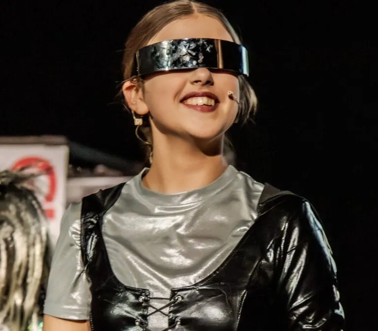 Child wearing metallic visor and futuristic costume on stage, smiling during a performance.