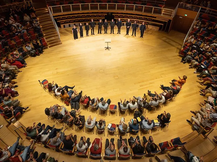 Classical musicians and a conductor arranged in a spiral formation on a wooden concert hall stage, viewed from above with audience seated in tiered balconies.