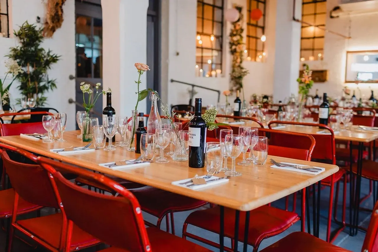 Long wooden dining table set for a formal event with red chairs, wine bottles, glassware, and floral centrepieces in a modern industrial space.