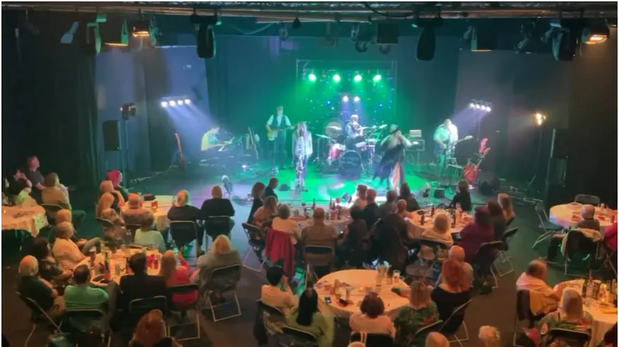 Audience seated at tables watches a live band performing on an illuminated stage with green and purple lighting.